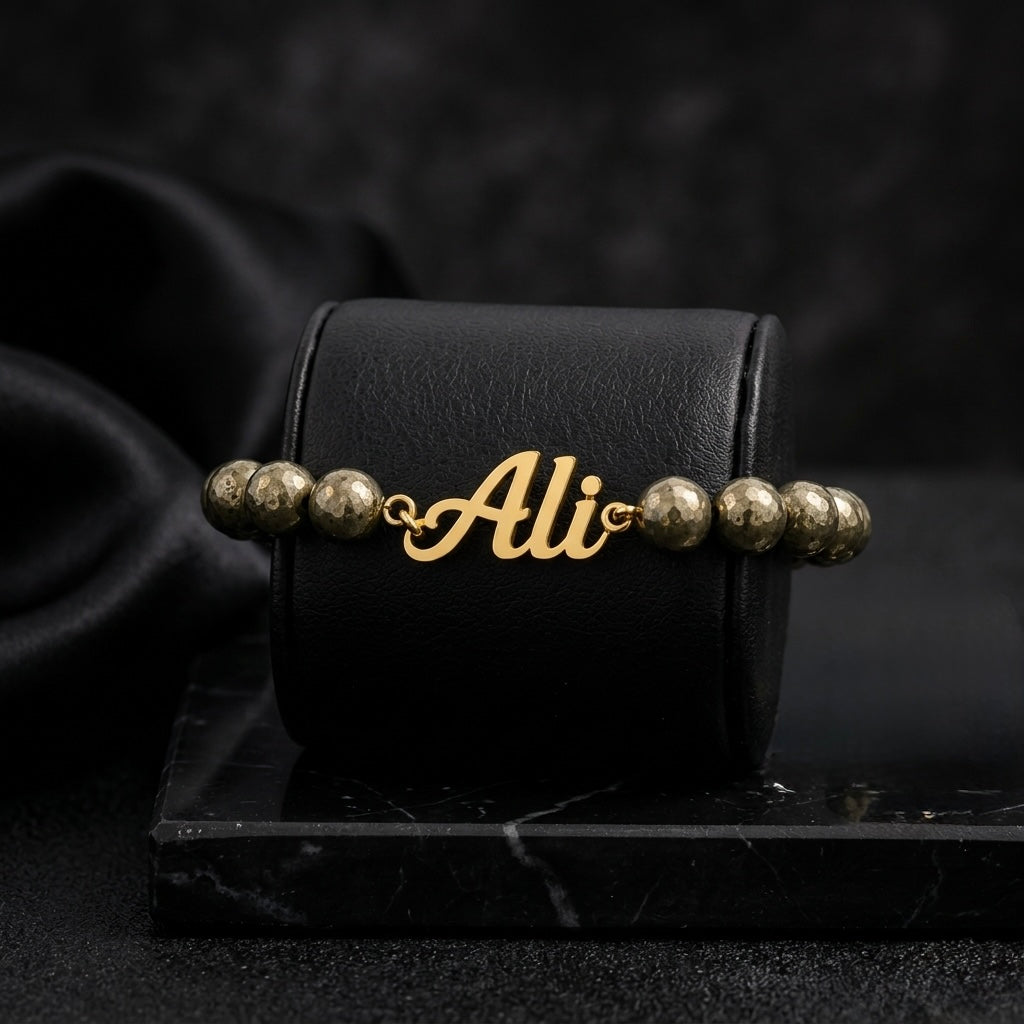 Gold bracelet with 'Ali' charm on a black box with 'Ali' branding, dark background