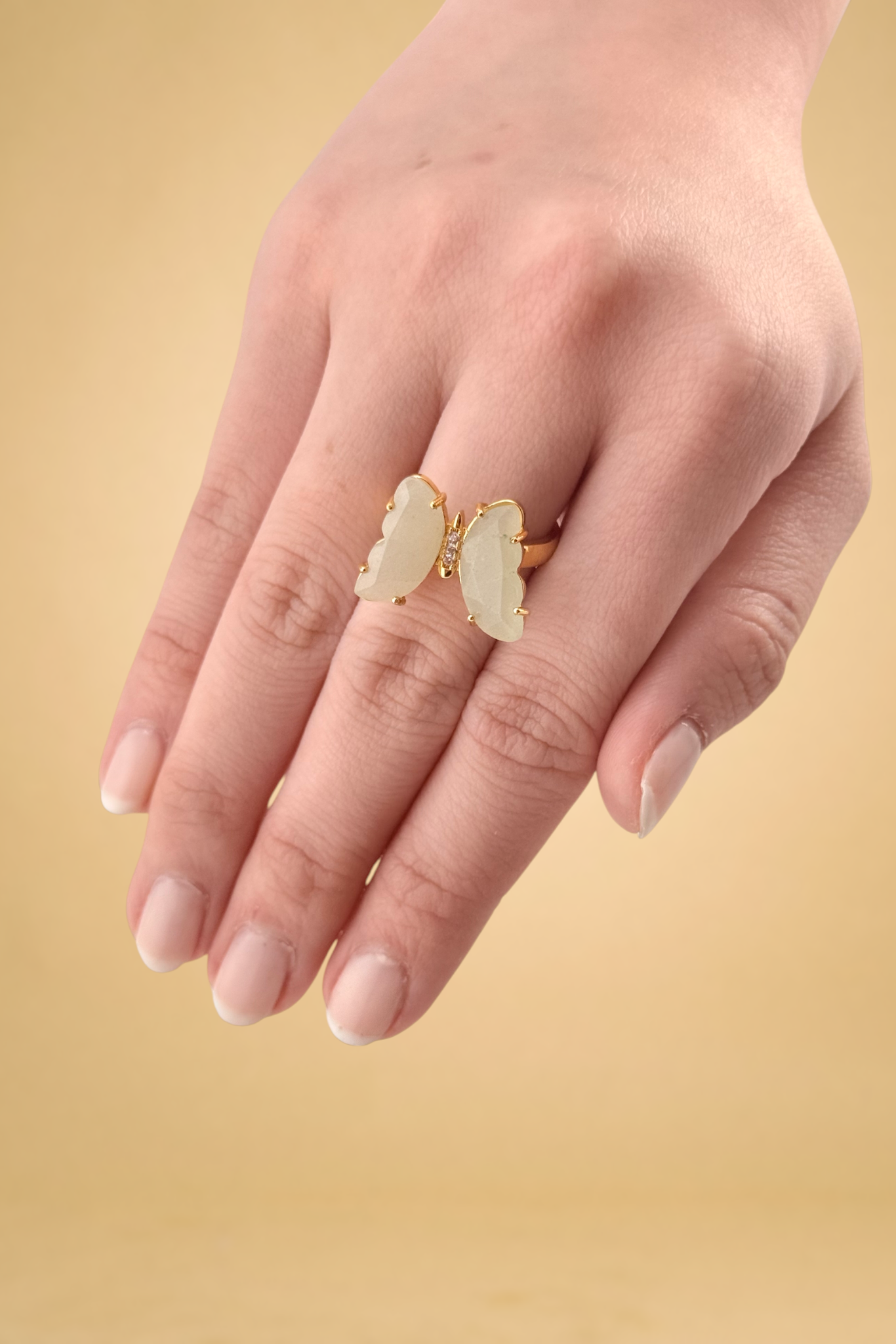Hand wearing a gold ring with a white stone on a beige background
