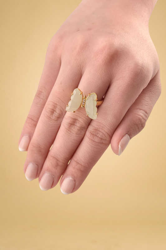 Hand wearing a gold ring with a white stone on a beige background