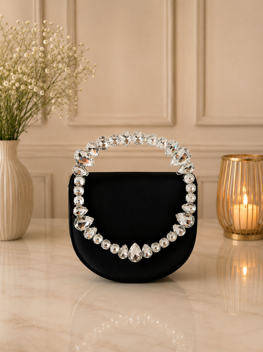 Black handbag with crystal handle on a beige surface, decorative vase, and candle in the background.
