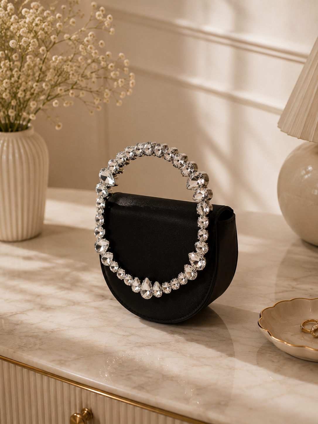 Black handbag with a decorative silver handle on a marble surface, with a vase and lamp in the background.