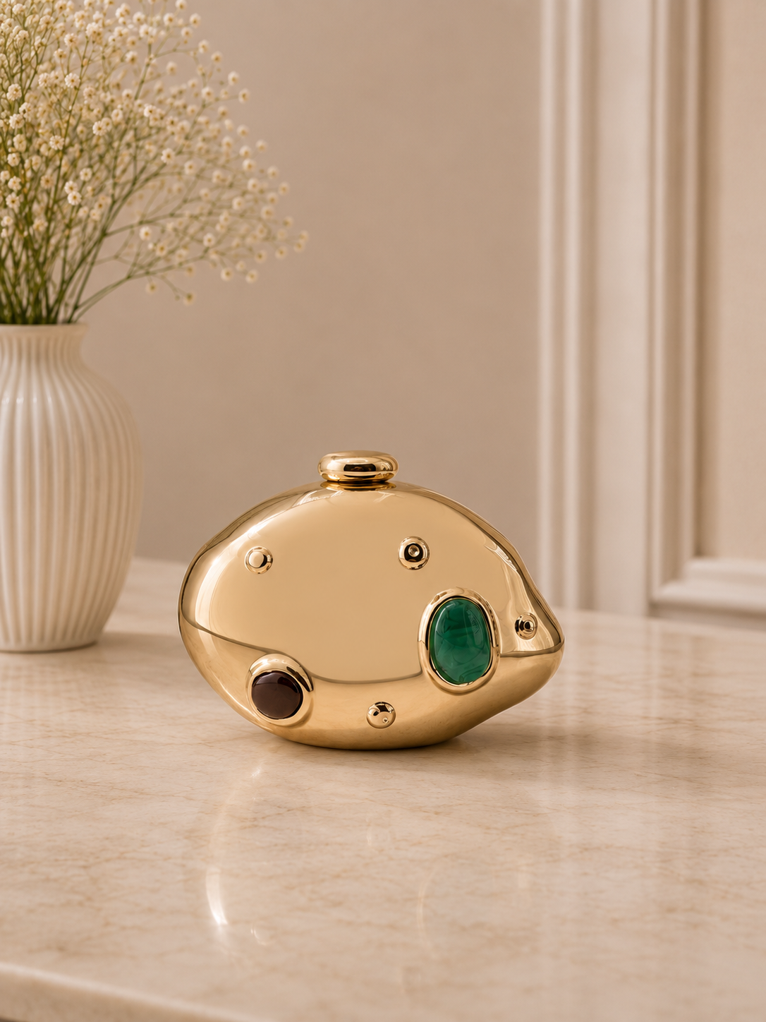Gold decorative object with a green gemstone on a marble surface, with a vase of flowers in the background.