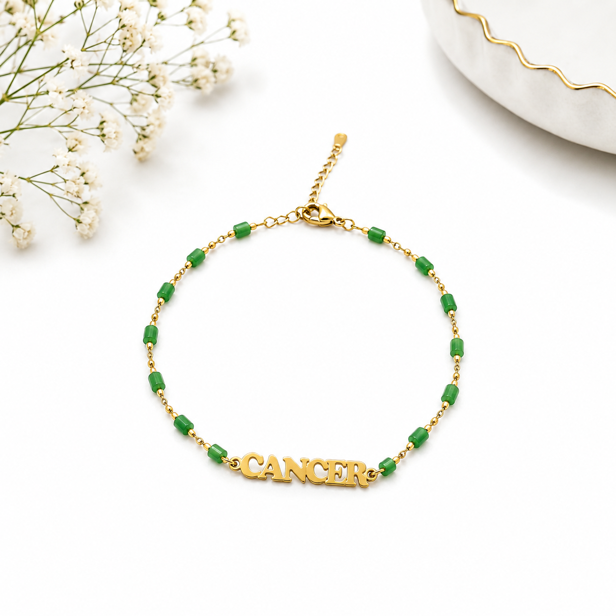 Gold bracelet with green beads spelling 'Cancer' on a white background with small flowers.