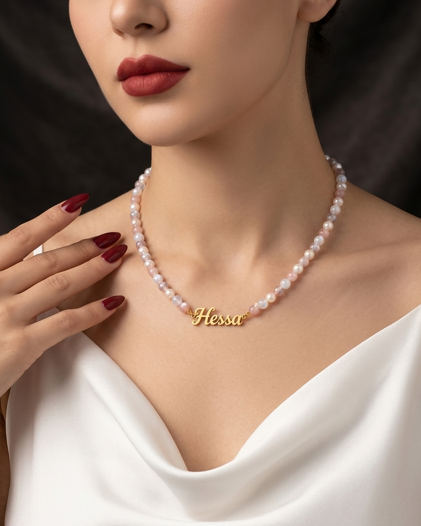 Woman wearing a pearl necklace with a gold nameplate, set against a dark background.