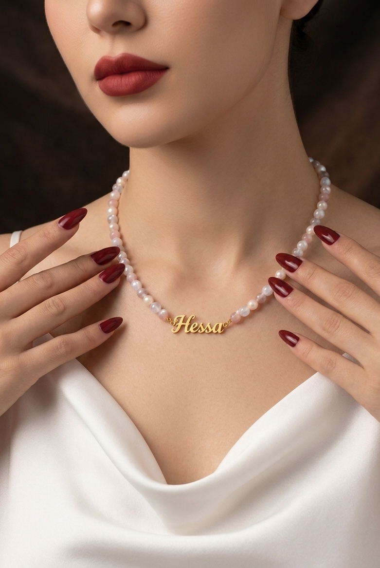 Woman wearing a pearl necklace with a gold nameplate, set against a dark background.