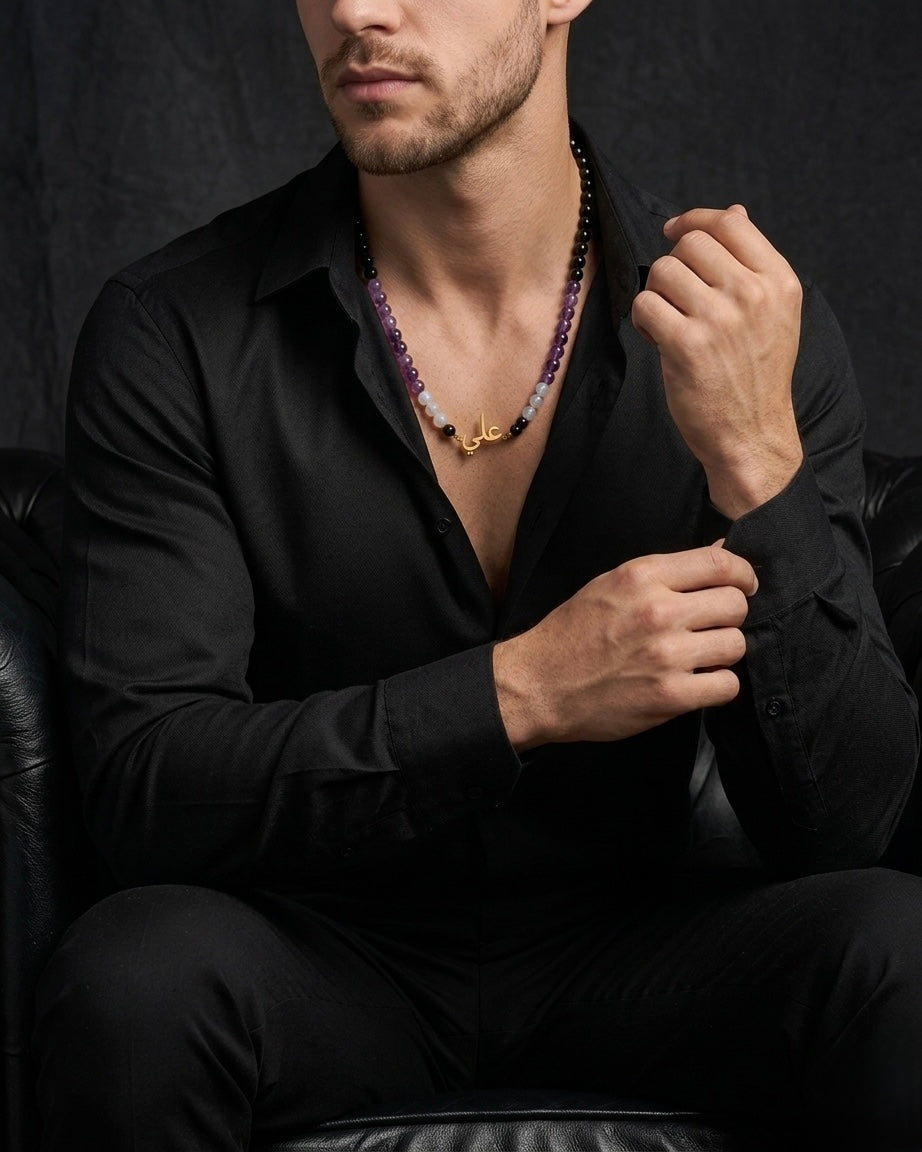 Man wearing a black shirt with a necklace, sitting against a dark background
