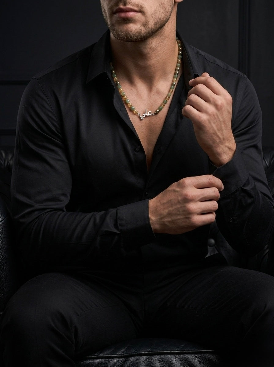 Man wearing a black shirt and dark pants with a necklace, sitting against a dark background.