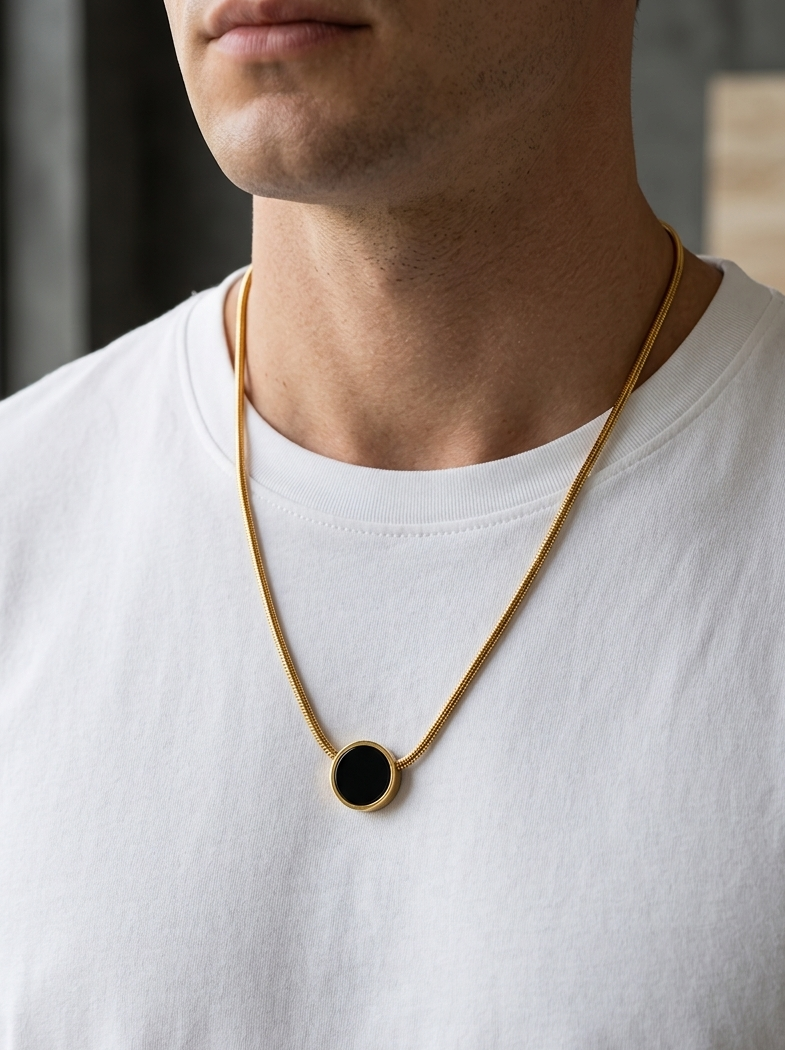 Man wearing a gold necklace with a black pendant on a white shirt.