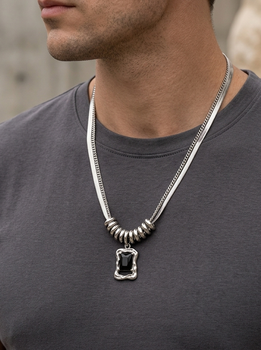 A close-up of a man wearing a silver-toned chain necklace with a rectangular pendant featuring a black center, styled with a dark gray t-shirt.