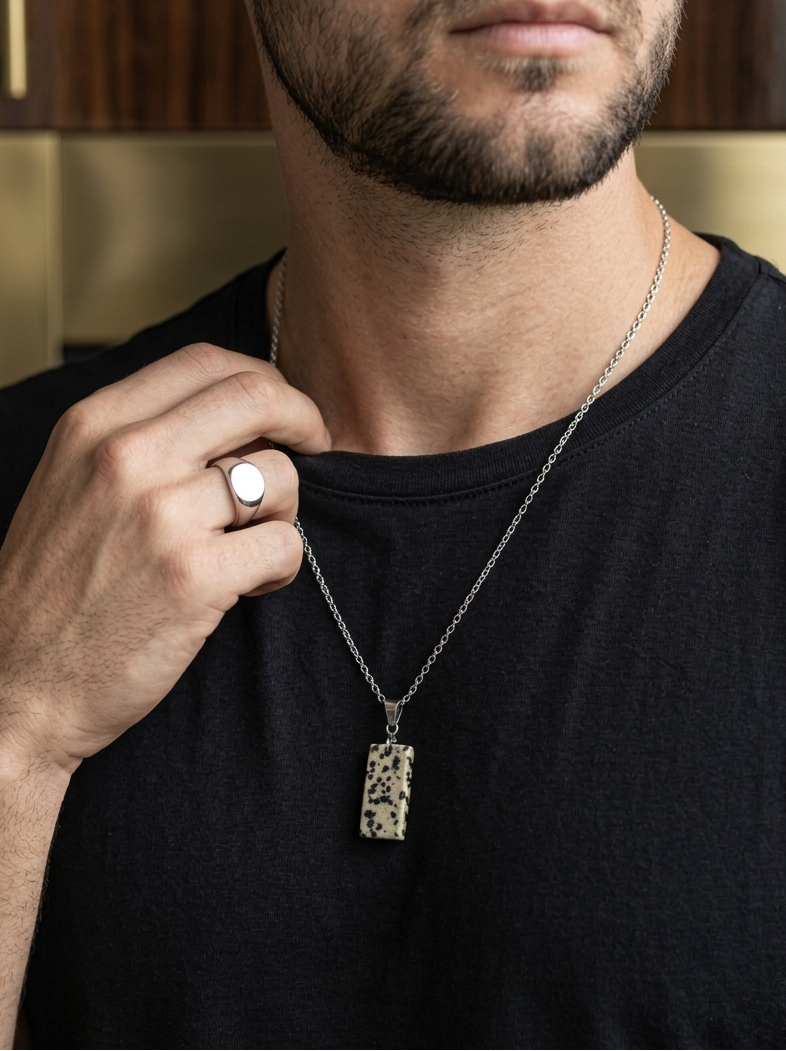 Man wearing a black shirt with a necklace and ring, against a blurred indoor background.