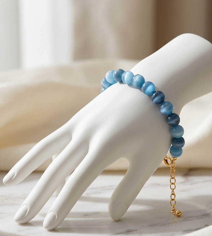 A beaded bracelet in shades of blue with a gold chain extender, displayed on a white mannequin hand over a marble surface.
