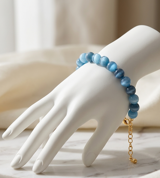 A beaded bracelet in shades of blue with a gold chain extender, displayed on a white mannequin hand over a marble surface.