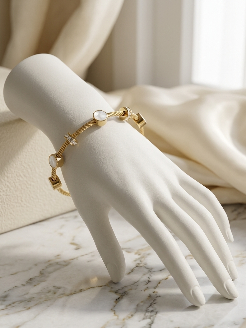 A gold bracelet with small geometric charms and a round white stone, displayed on a white mannequin hand over a marble surface with cream fabric in the background.