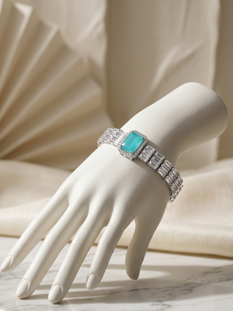 A silver-toned bracelet with a rectangular turquoise-blue center stone and pavé-set clear stones, displayed on a white mannequin hand over a marble surface with draped cream fabric in the background.