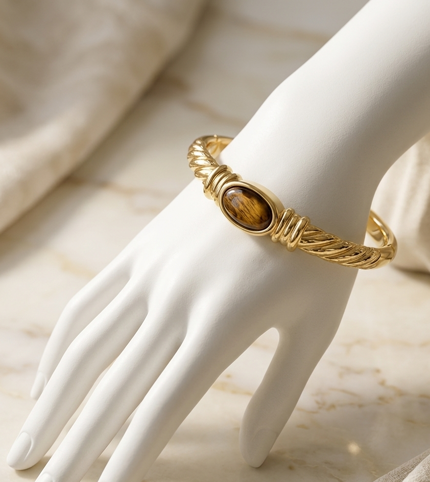 A gold-toned twisted rope bangle bracelet with an oval tiger's eye stone, displayed on a white mannequin hand over a beige marble surface.