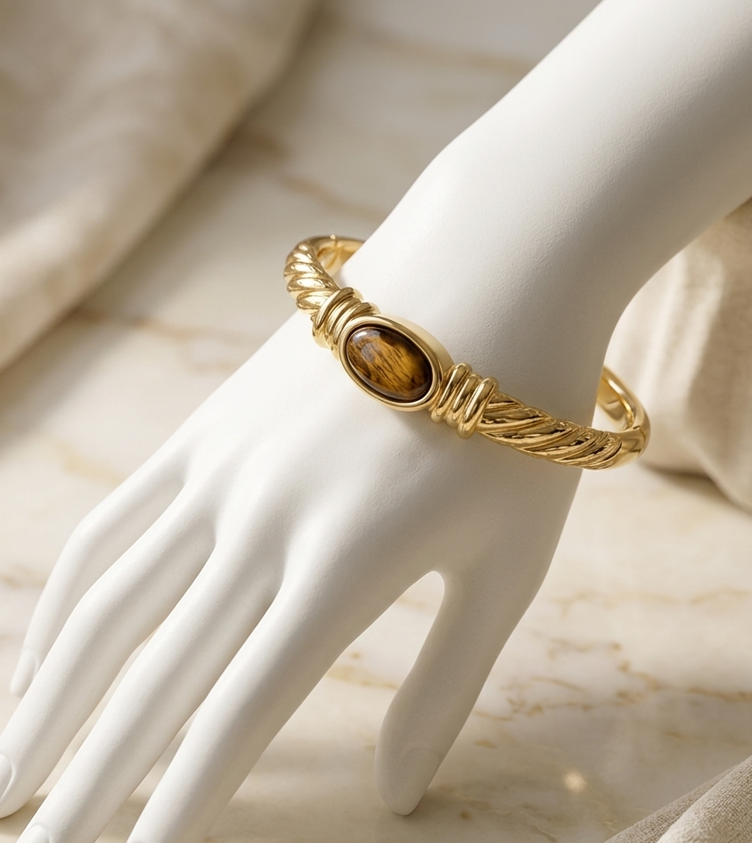 A gold-toned twisted rope bangle bracelet with an oval tiger's eye stone, displayed on a white mannequin hand over a beige marble surface.