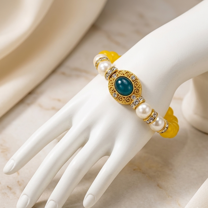 A gold-toned bracelet with a central turquoise-blue oval stone, white pearl-like beads, and yellow translucent accents, displayed on a white mannequin hand against a soft cream fabric and marble surface.