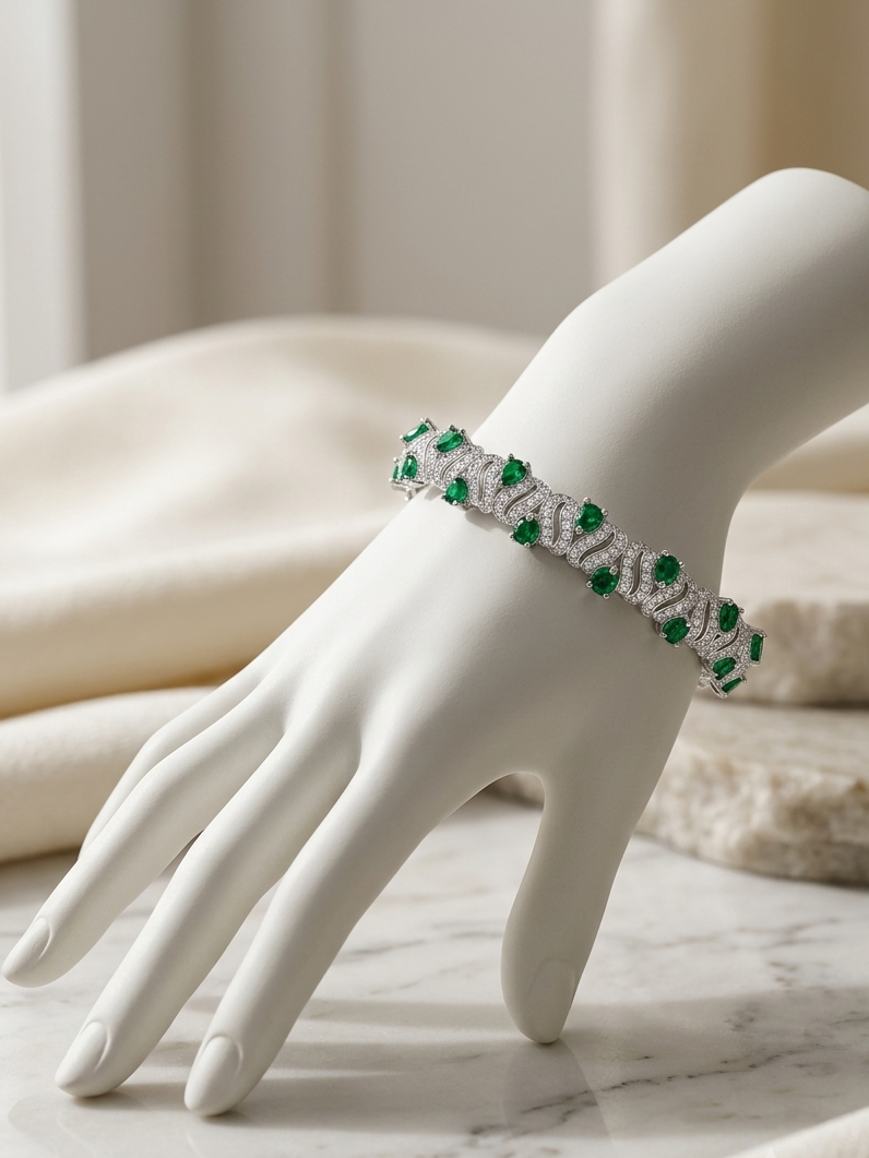 A white gemstone and emerald bracelet displayed on a white mannequin hand, resting on a marble surface with soft neutral fabric in the background.