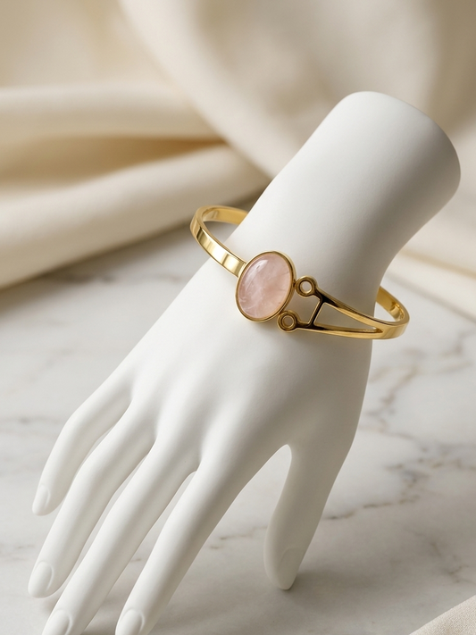 A gold bangle bracelet with an oval pink stone centerpiece, displayed on a white mannequin hand over a light marble surface with draped cream fabric in the background.