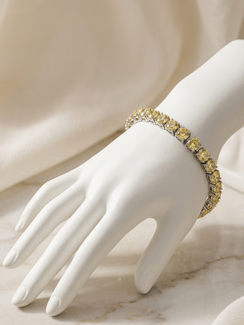 A yellow stone tennis bracelet with silver-toned metal settings displayed on a white mannequin hand, set against a draped cream fabric and light marble surface.