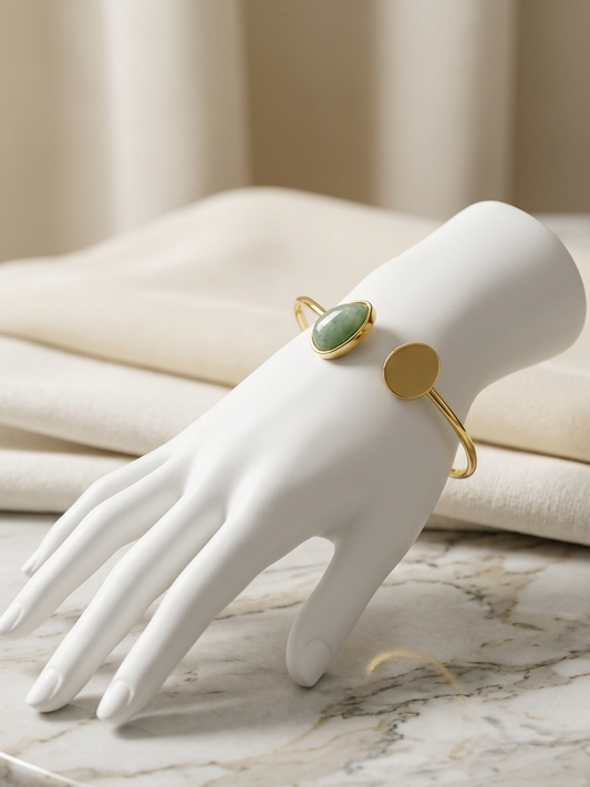 A gold cuff bracelet with a green oval stone and a flat gold disc, displayed on a white mannequin hand over a marble surface with folded cream fabric in the background.