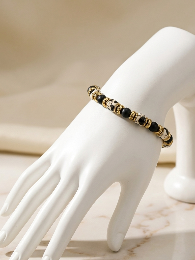 A gold and black beaded bracelet displayed on a white mannequin hand against a soft beige background.
