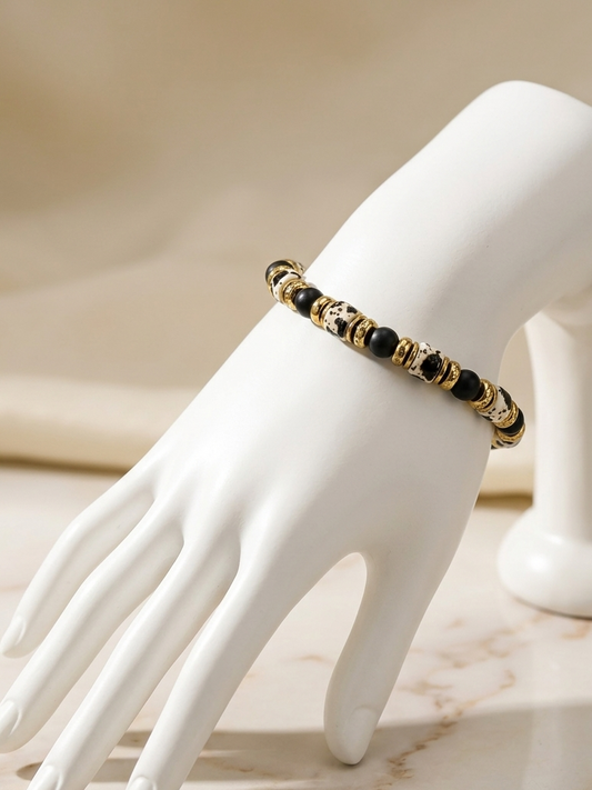 A gold and black beaded bracelet displayed on a white mannequin hand against a soft beige background.