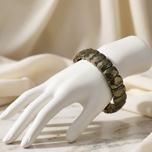A dark metallic segmented bracelet displayed on a white mannequin hand, set against a soft cream fabric backdrop on a light marble surface.