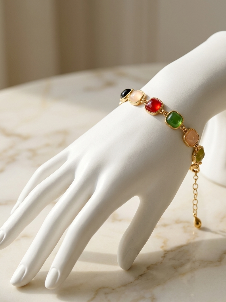 A gold chain bracelet with colorful gemstone-like beads in red, green, black, and gold-toned oval details, displayed on a white mannequin hand over a light marble surface.