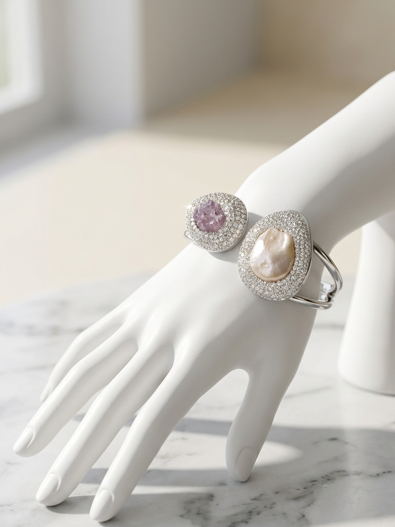A white jewelry display hand wearing two silver rings. One ring features a round pink gemstone surrounded by clear stones, and the other features an oval cream-colored pearl or shell centerpiece with a halo of clear stones.