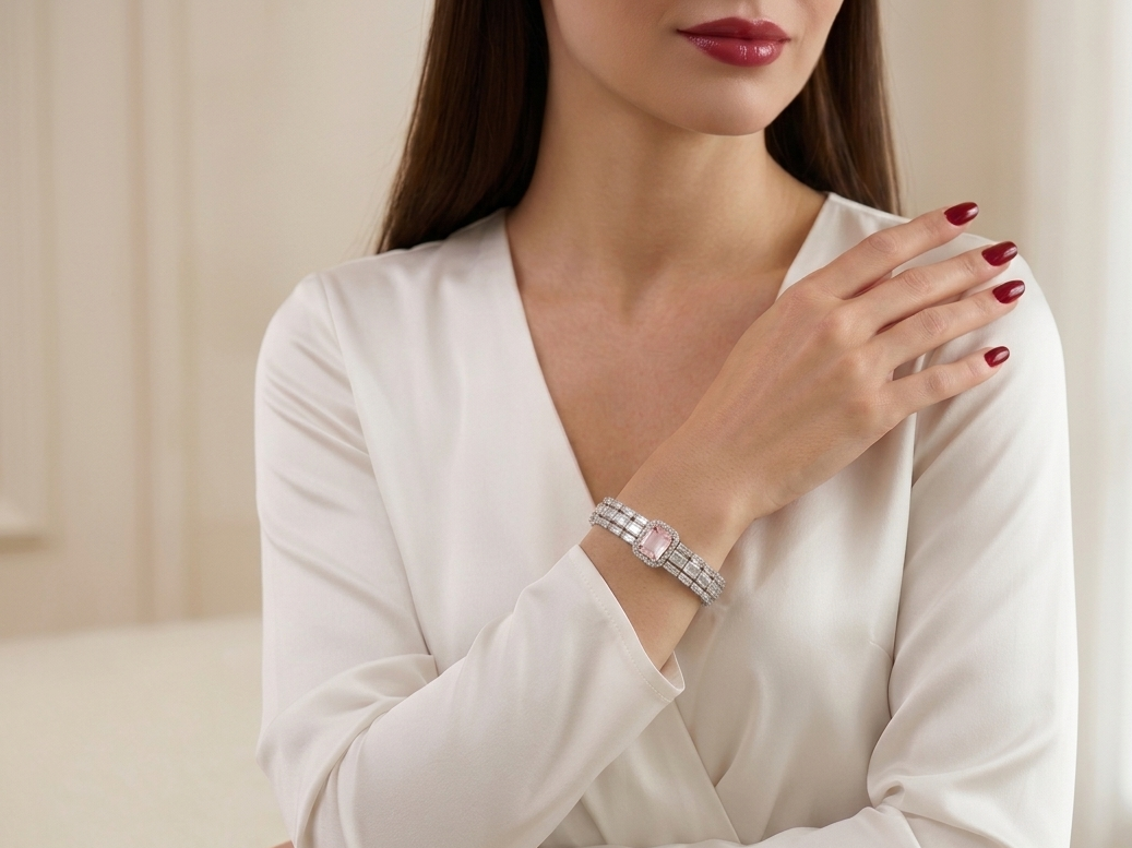 A woman wearing a white long-sleeve top and red nail polish, showcasing a silver-toned diamond-embellished bracelet on her wrist in a softly lit indoor setting.