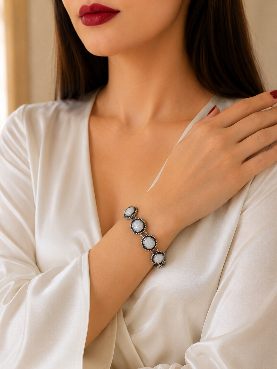 A woman wearing a white satin blouse and dark red lipstick, showcasing a silver-toned bracelet with five oval white stones on her wrist.
