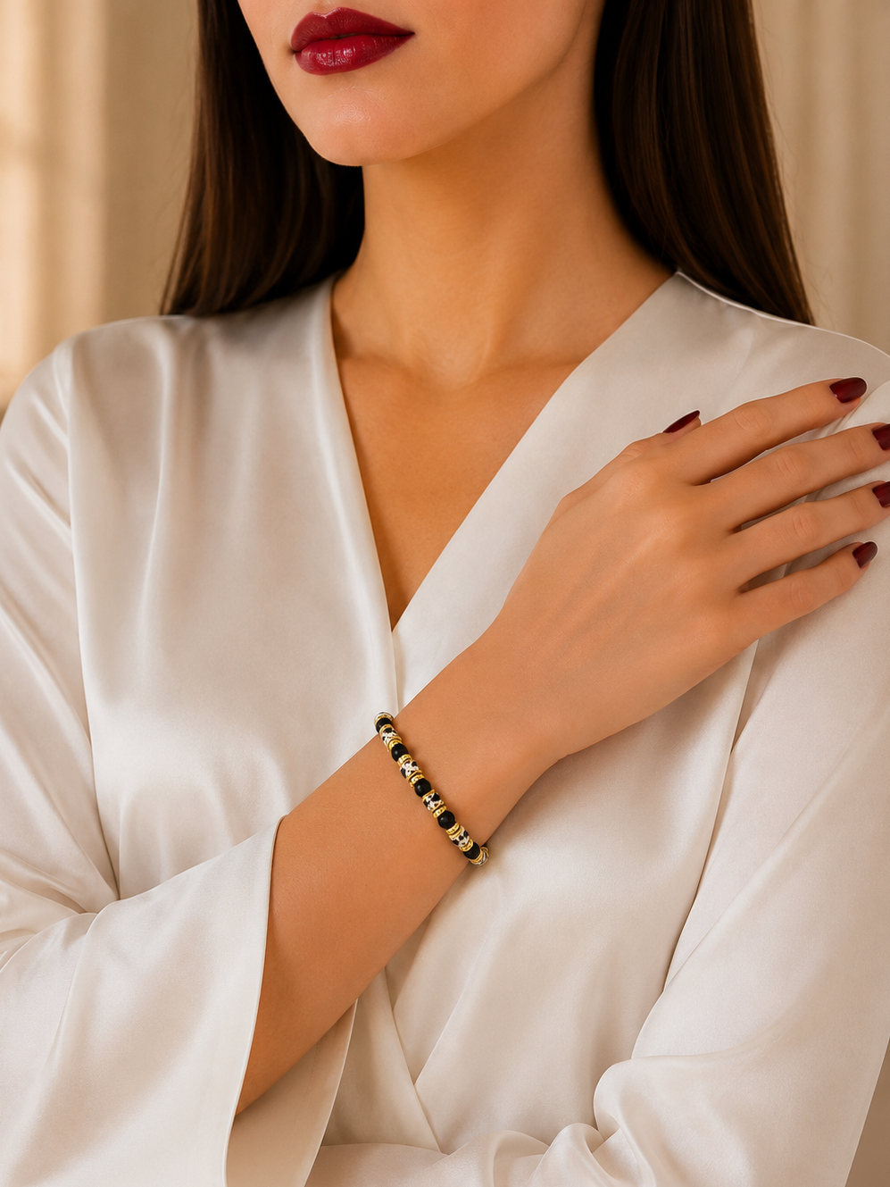 A close-up of a woman wearing a delicate beaded bracelet on her wrist, dressed in a white satin robe. The bracelet features alternating black and gold beads.