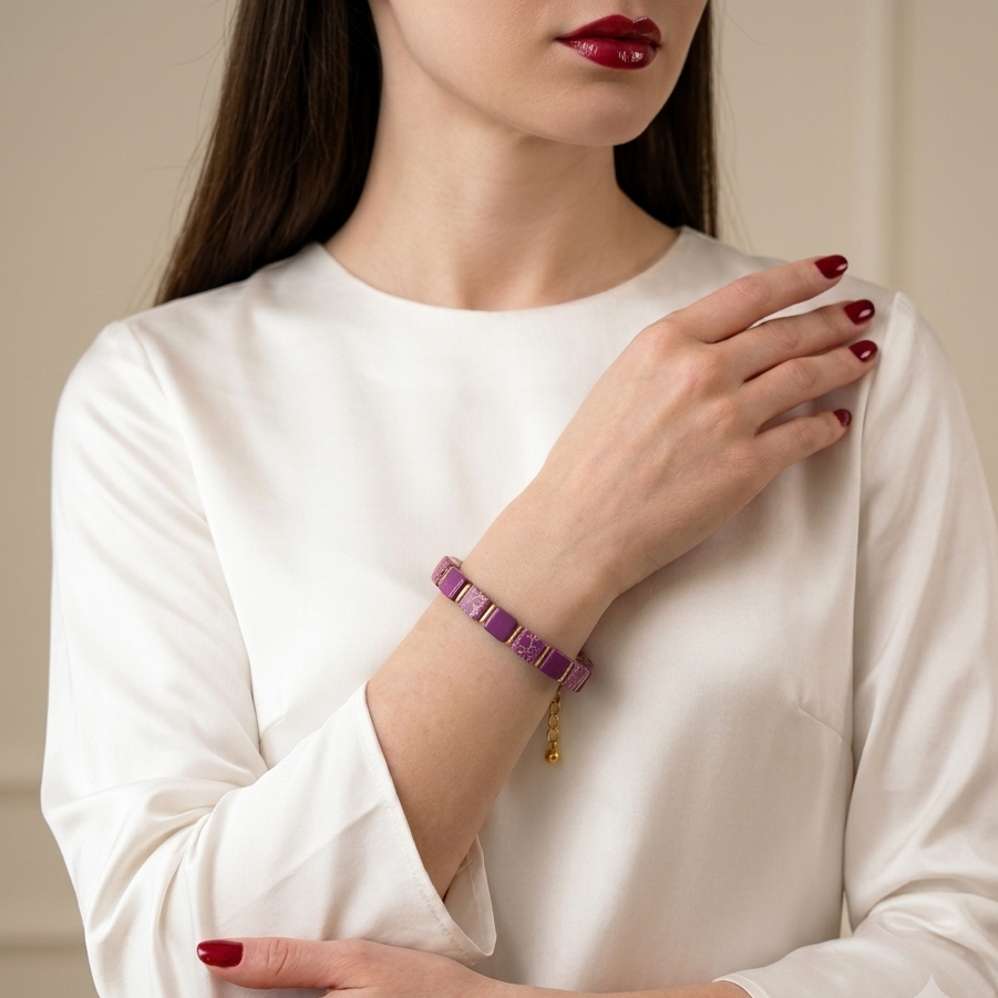 A woman wearing a purple beaded bracelet with a small gold charm, paired with a white long-sleeve blouse. The photo is cropped from the lips down, showing her hands and arms against a soft beige background.