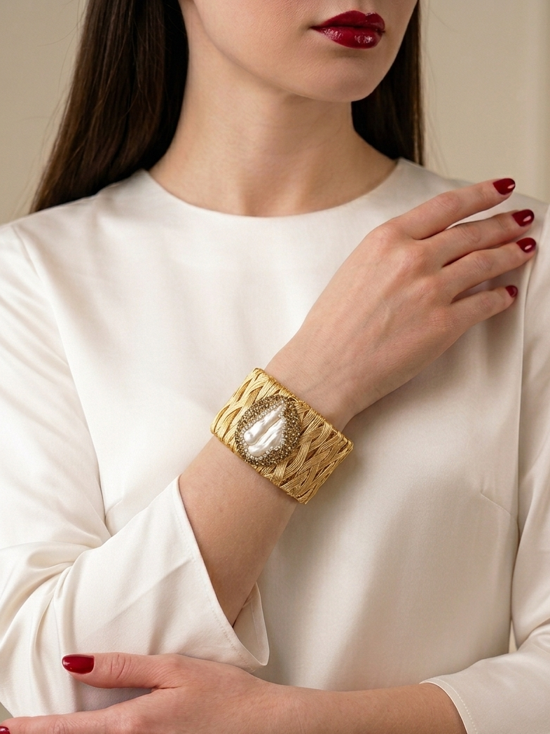 A close-up of a woman wearing a wide gold cuff bracelet with a textured woven pattern and a central oval crystal-like detail, paired with a white blouse and red nail polish.