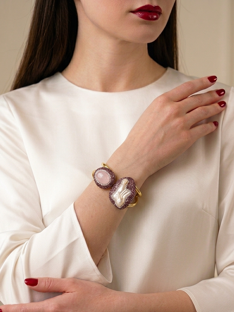 A close-up of a woman wearing a gold-toned ornate cuff bracelet with a central amber stone and a white pearl, paired with a white satin blouse.