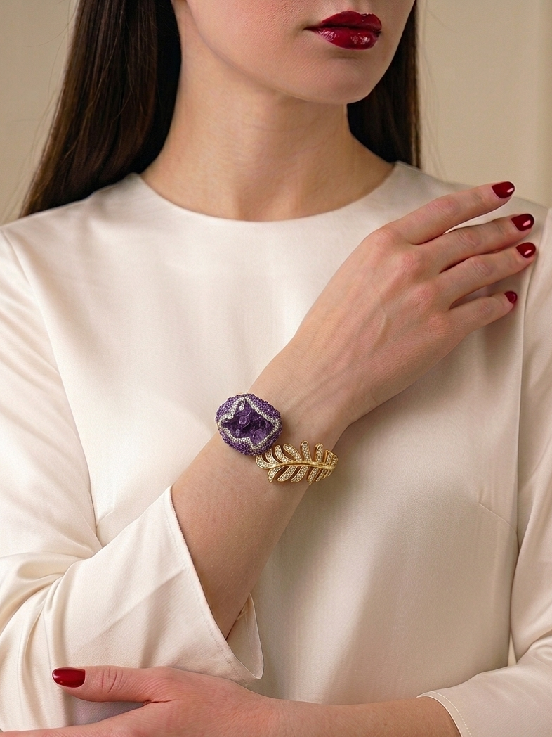 A woman wearing a cream-colored satin blouse and red nail polish, showcasing a gold leaf bracelet with a purple amethyst crystal cabochon on her wrist.