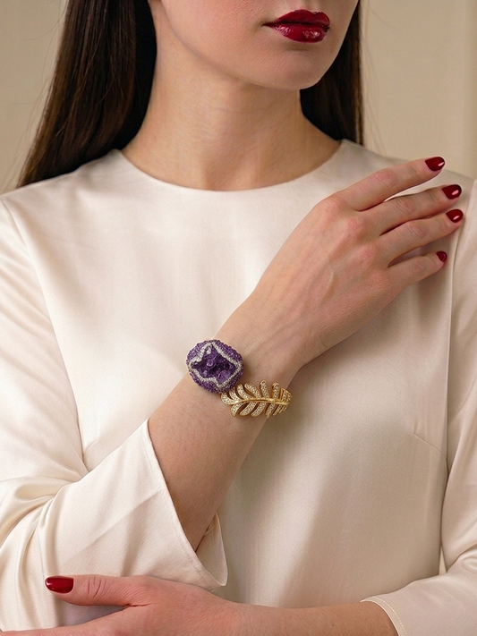 A woman wearing a cream-colored satin blouse and red nail polish, showcasing a gold leaf bracelet with a purple amethyst crystal cabochon on her wrist.