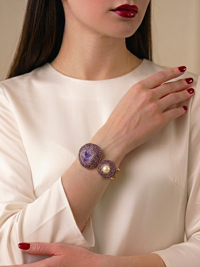 A close-up of a woman wearing a purple and gold cuff bracelet with a pearl accent, paired with a cream satin blouse and red nail polish.