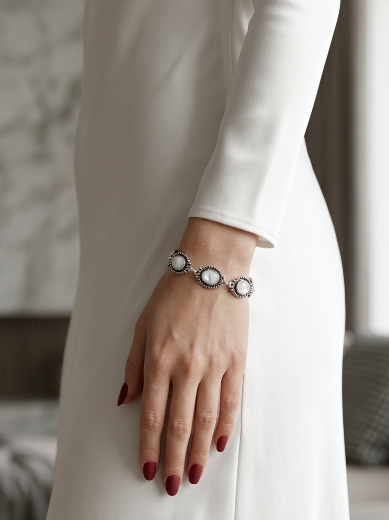 A close-up of a person wearing a silver-toned bracelet with three oval white stones, shown against a white long-sleeve dress in a softly lit indoor setting.