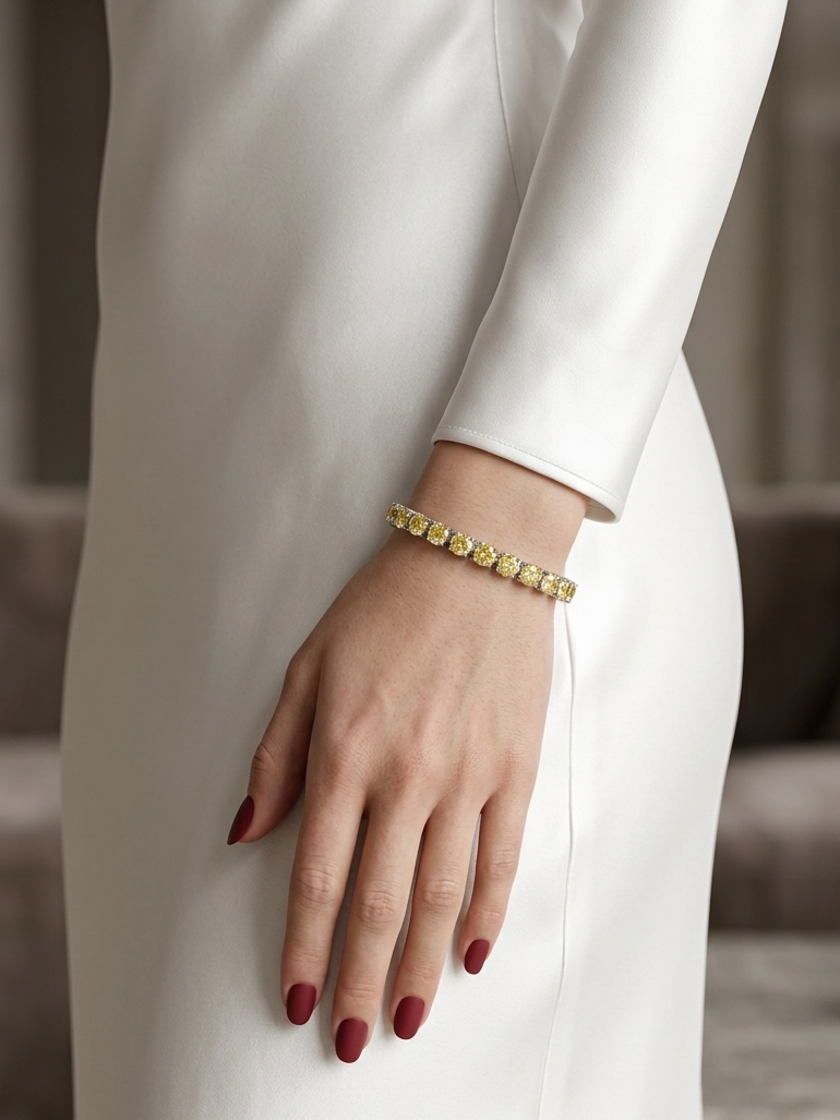 A close-up of a model's hand wearing a delicate gold bracelet with small, round-cut clear stones, styled with a long-sleeved white dress and dark red manicured nails.