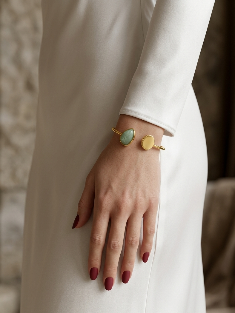 A close-up of a person wearing a gold cuff bracelet with two oval stones, one light green and one matte gold, styled with a white long-sleeve dress.