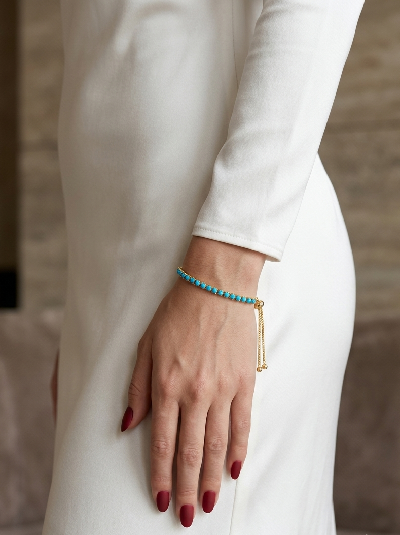 A close-up of a woman's hand and wrist wearing a delicate turquoise beaded bracelet with a gold bar tassel, styled with a white long-sleeve dress.