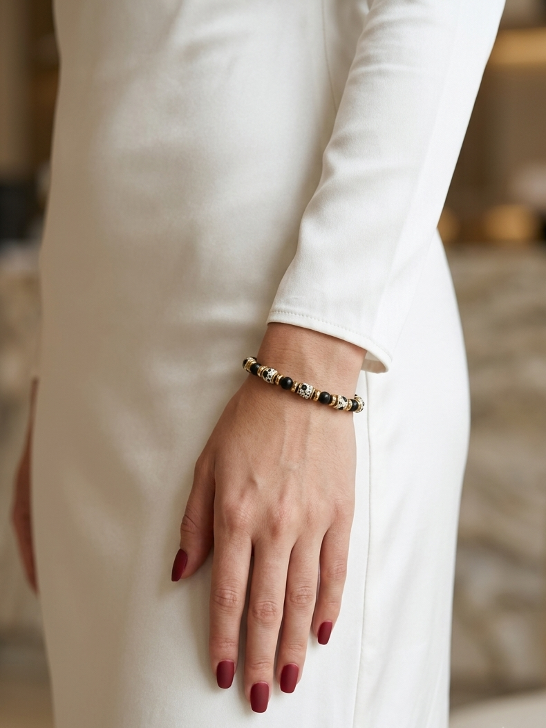 A close-up of a person wearing a delicate beaded bracelet on their wrist, dressed in a long-sleeved white dress. The bracelet features alternating dark brown and metallic gold beads.