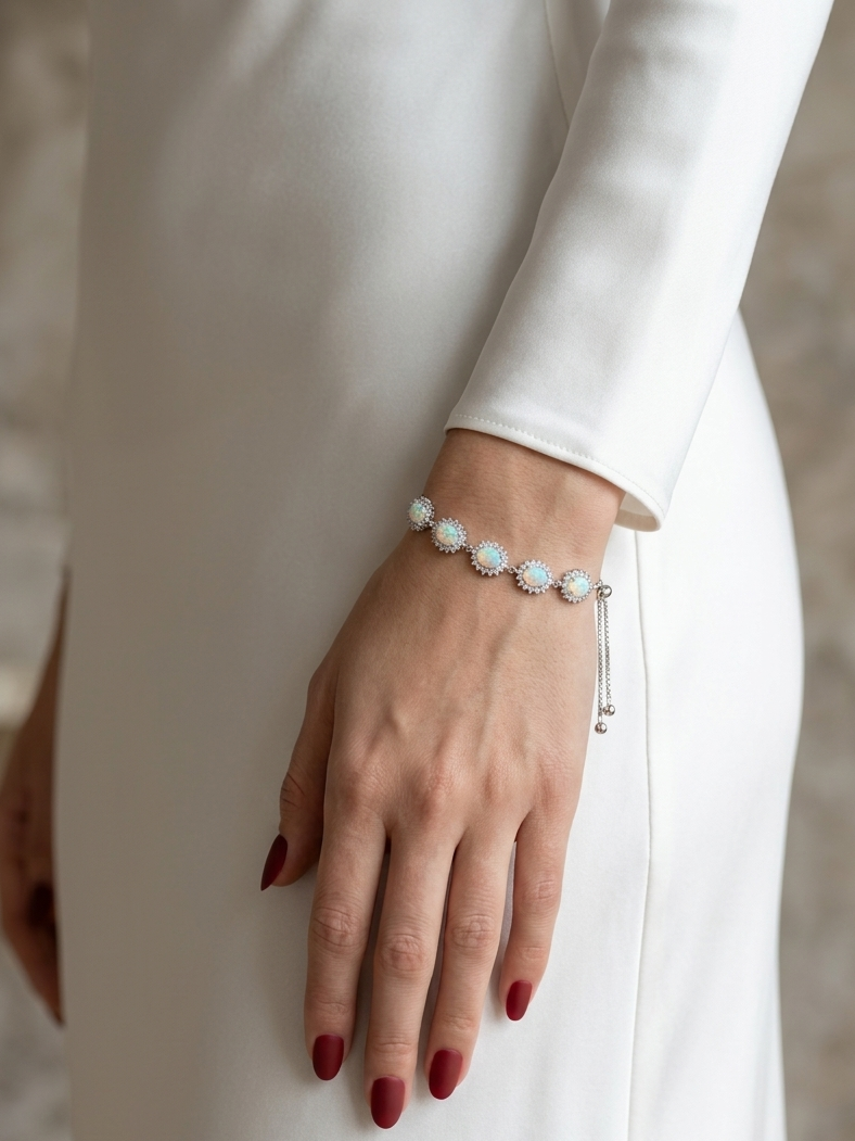 A close-up of a woman's hand wearing a delicate silver bracelet adorned with small white pearls and clear crystals, styled with a white long-sleeve dress.