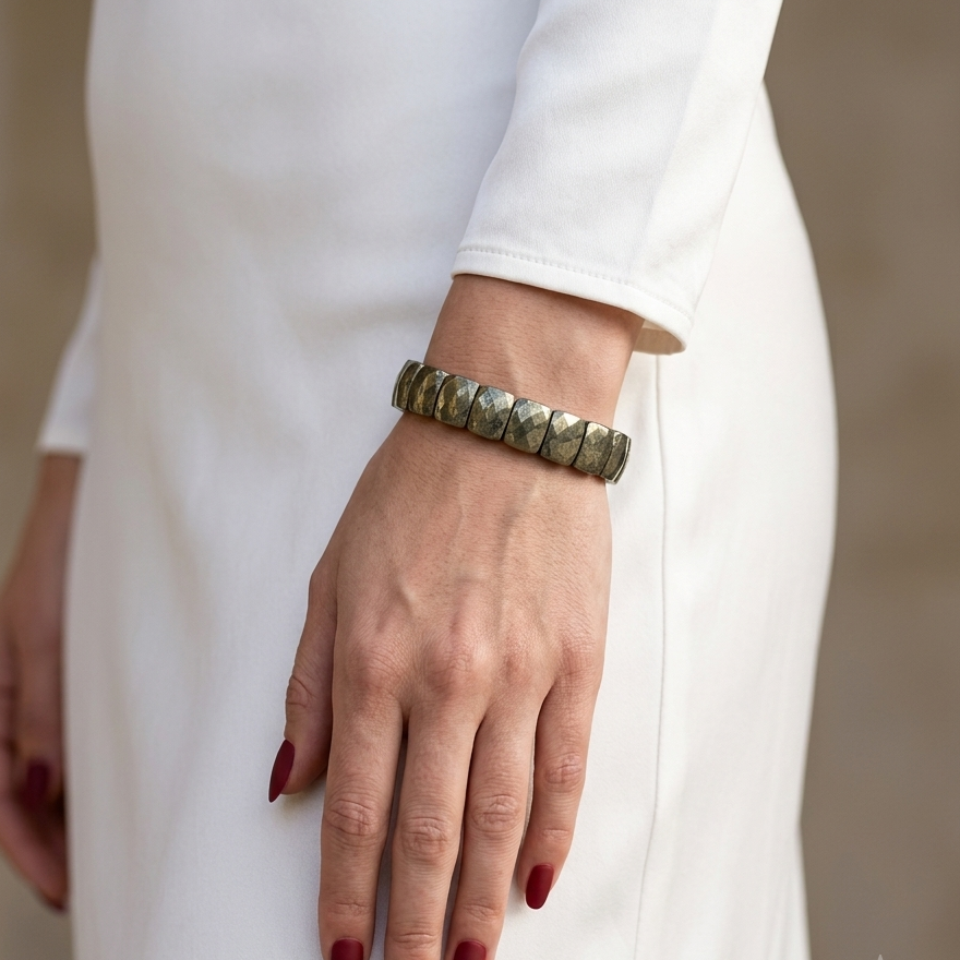 A close-up of a person wearing a chunky metallic cuff bracelet with a textured, hammered finish. The person is wearing a white long-sleeve dress, and the background is a soft, neutral beige wall.