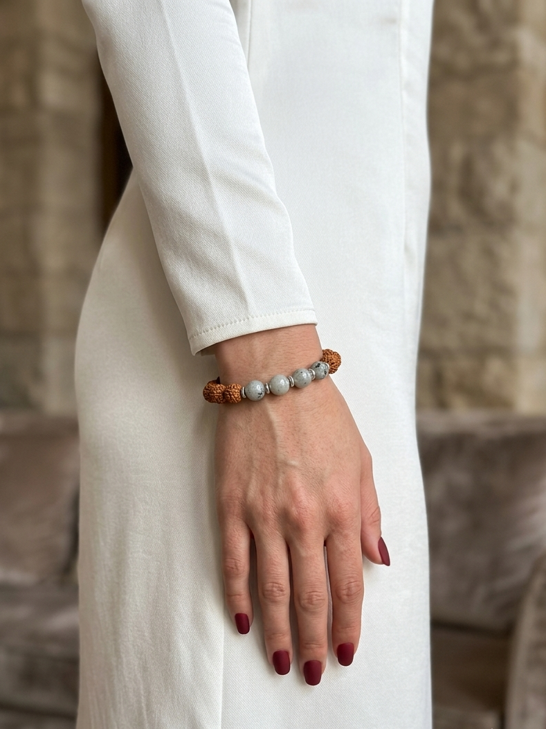 A close-up of a person's hand and forearm wearing a beaded bracelet with brown wooden beads and light gray stone beads, while wearing a long white dress in an indoor setting.