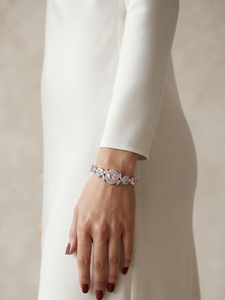 A close-up of a model's hand wearing a delicate bracelet with pink and clear gemstones, styled with a long-sleeved white dress against a soft beige background.