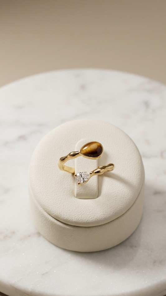 A delicate gold ring with a small clear heart-shaped gemstone and a larger polished gold bead, displayed on a cream-colored ring stand atop a round white marble tray.