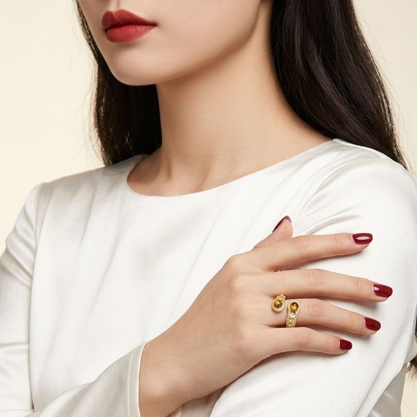 A close-up of a woman wearing a white satin long-sleeve top and a gold ring with oval-cut yellow gemstones on her hand, set against a warm cream background.